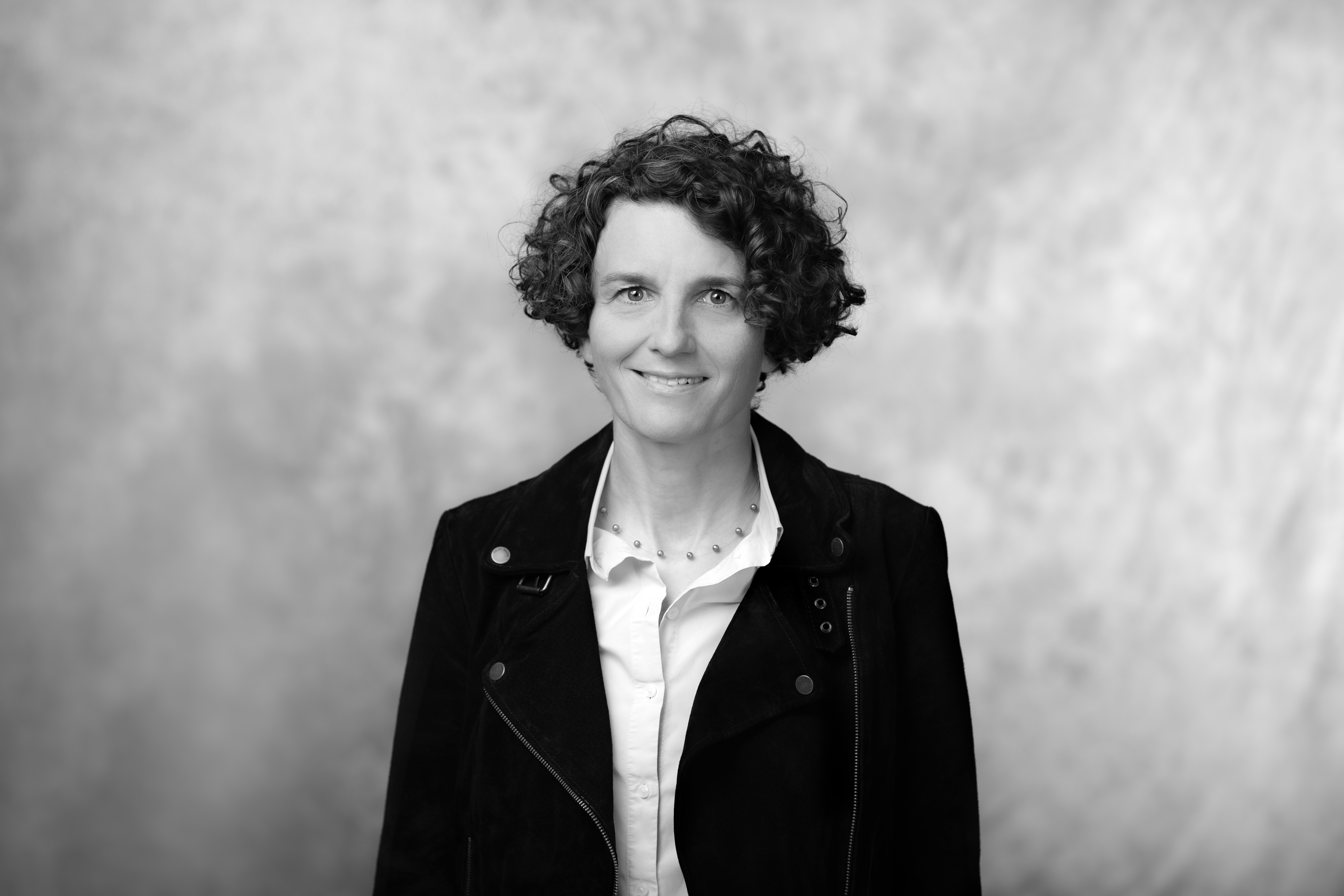 czafrno biały portret kobiety dojrzałej, uśmiechniętej, w czarnej kurtce i z kręconymi włosami długości do ucha