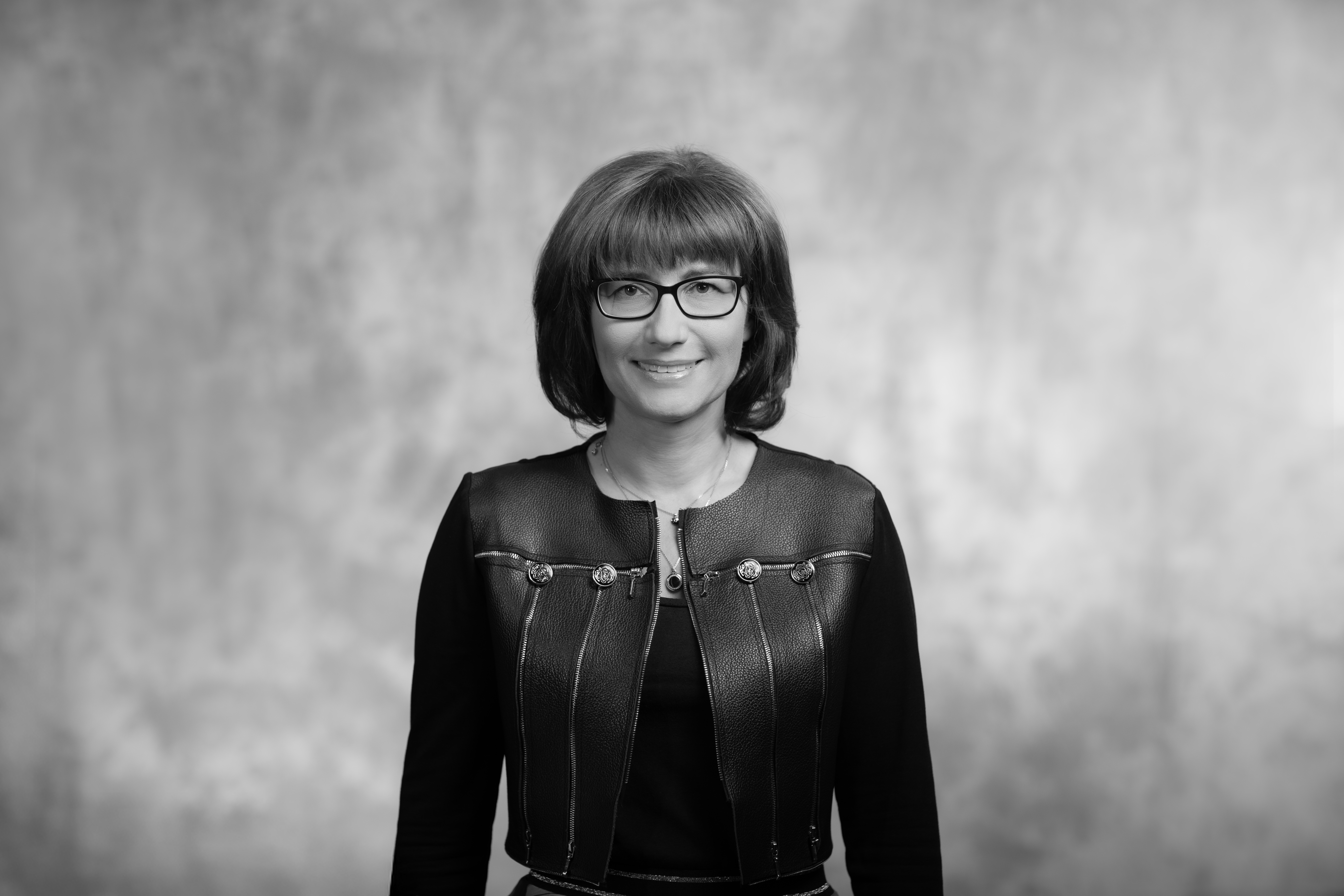 portet kobiety delikatnie się uśmiechającej, w okularach, stoi na tle ściany szarej