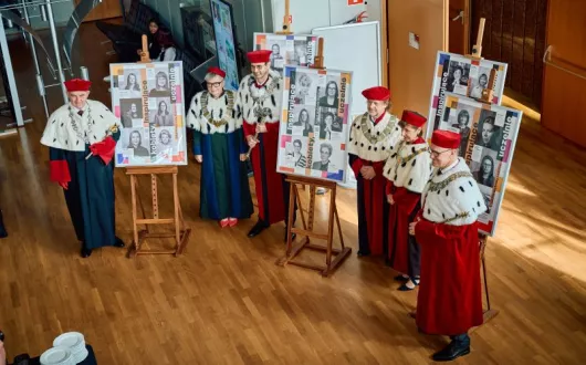 ujęcie z góry, 6 rektorów w oficjalnych strojach stoi w słonecznej sali między sztalugami