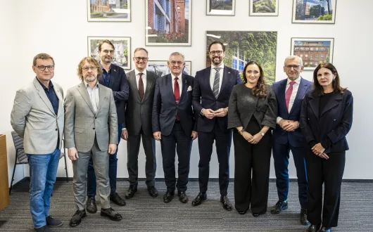 dziewięcioro osób elegancko ubranych stoi na tle jasnej ściany z oprawionymi zdjęciami