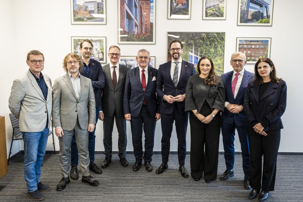 dziewięcioro osób elegancko ubranych stoi na tle jasnej ściany z oprawionymi zdjęciami